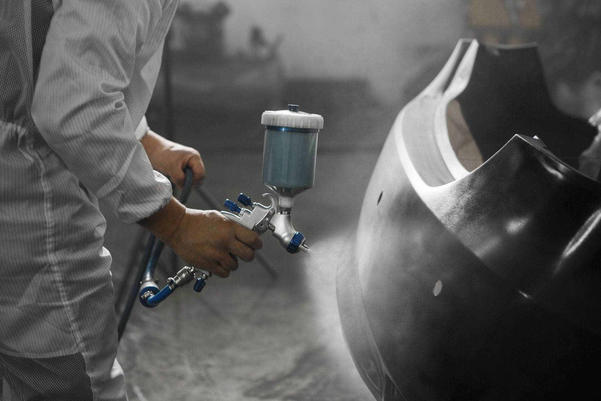 Une personne en combinaison de protection peint à la bombe un pare-chocs de voiture noir avec un pistolet à peinture bleu.