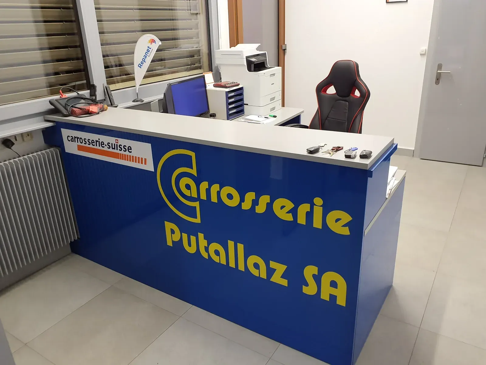 Un comptoir d'accueil bleu avec l'inscription jaune « Carrosserie Putallaz SA » dans un petit bureau.