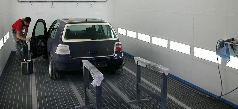 Une personne en chemise rouge travaille sur une voiture sombre à l'intérieur d'une cabine de peinture blanche.