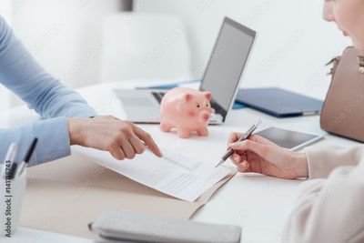 Zwei Personen sitzen an einem Schreibtisch mit einem Laptop und einem rosa Sparschwein und prüfen Unterlagen, was auf eine finanzielle Diskussion hindeutet.