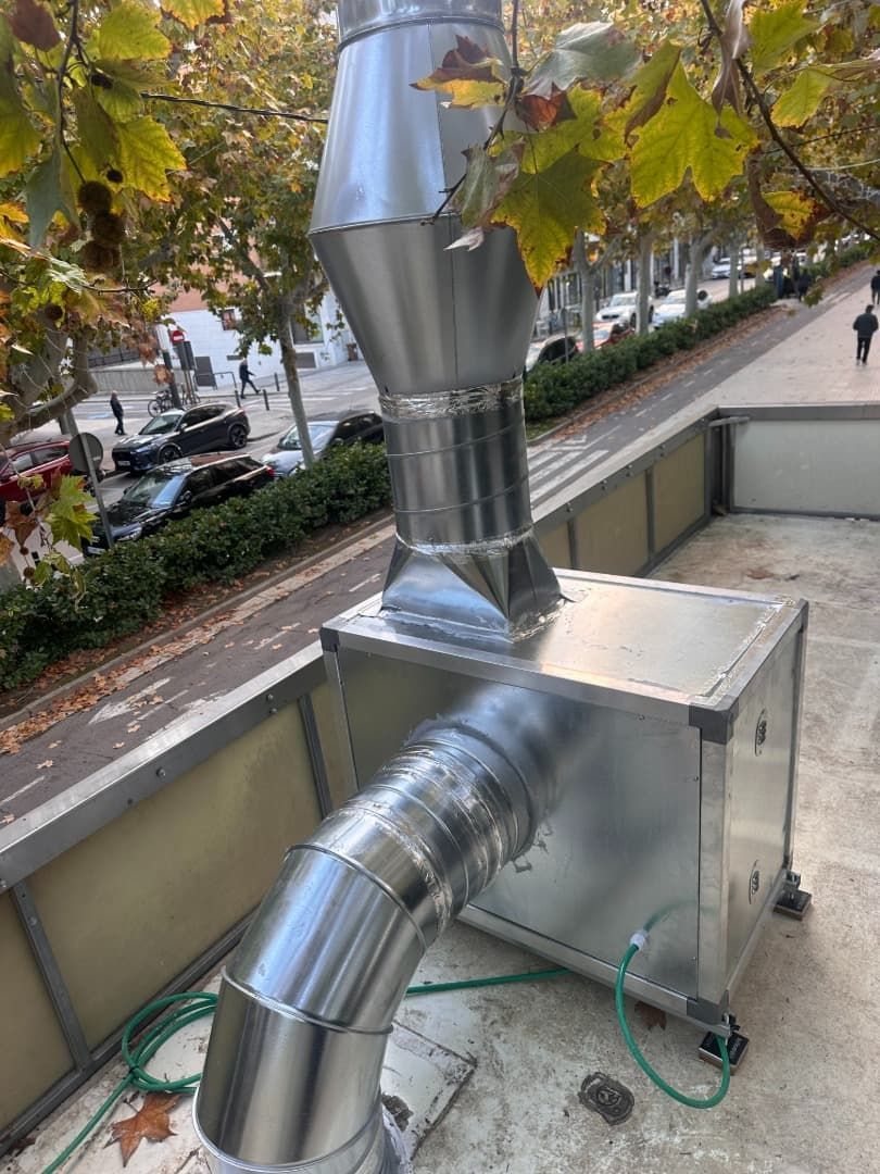 Sistema de ventilación de metal plateado en una azotea con un tubo curvo y una chimenea más alta, calle al fondo.