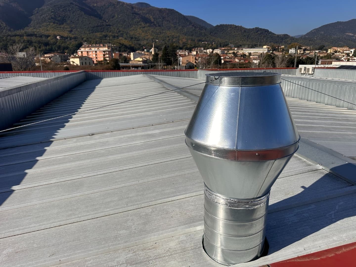 Rejilla de ventilación metálica sobre un tejado de chapa ondulada con una ciudad y montañas al fondo.