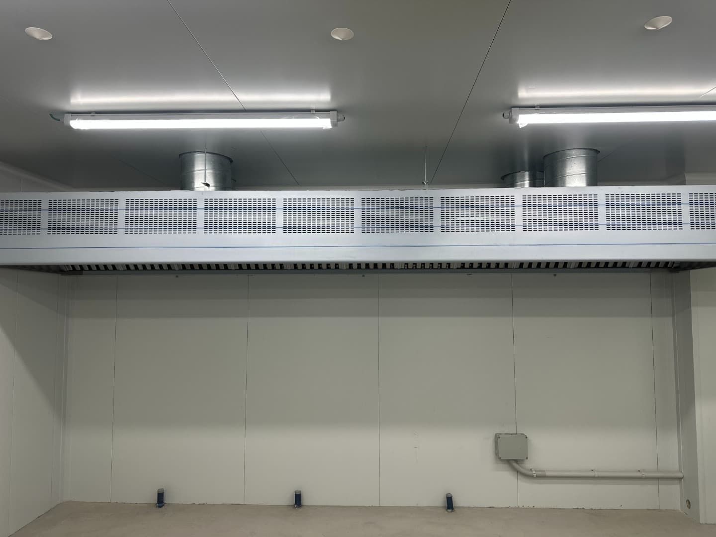 Un sistema de ventilación sobre una habitación de paredes blancas con piso gris y luces fluorescentes.