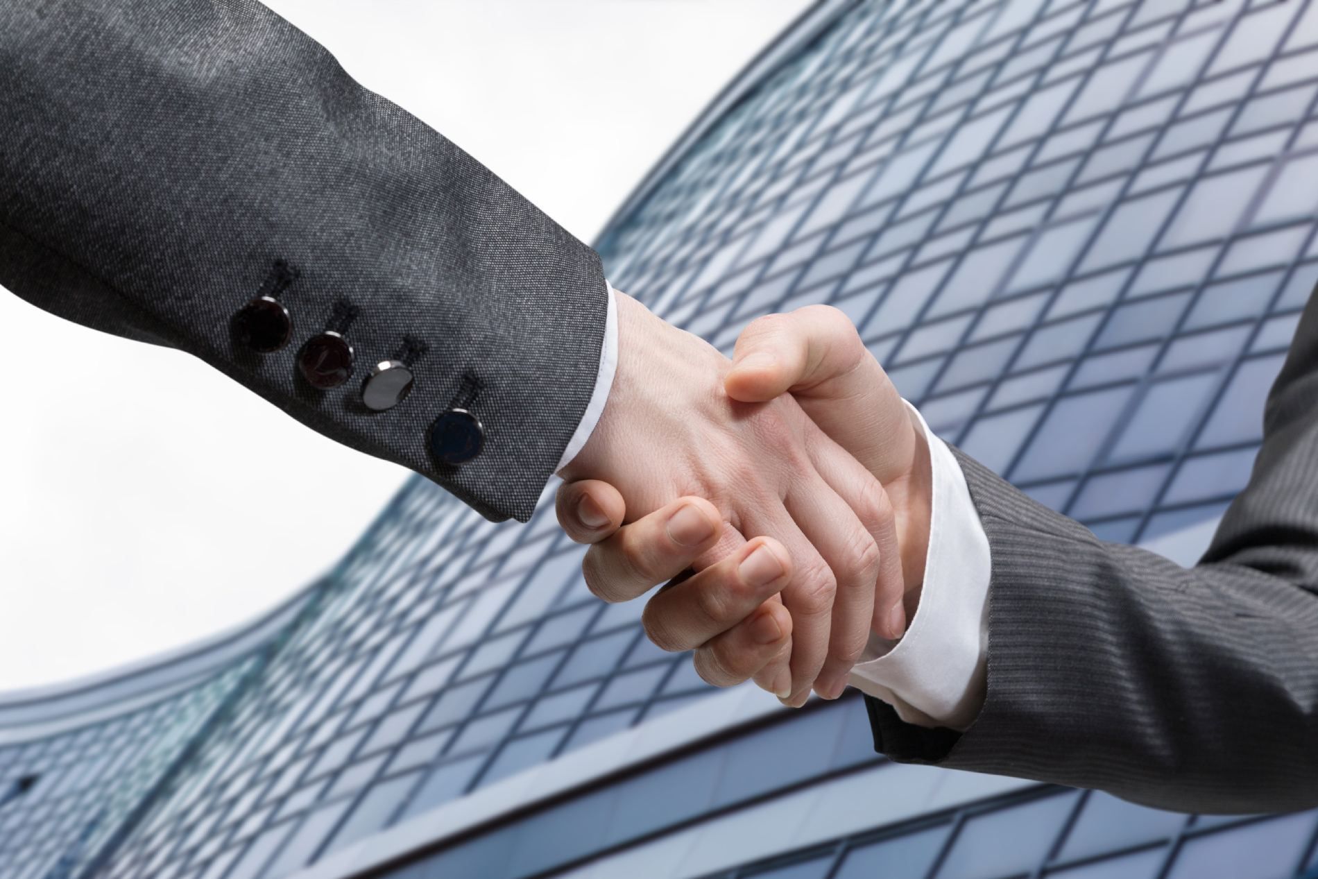 Dos personas con traje de negocios dándose la mano frente a un moderno edificio de oficinas de cristal.