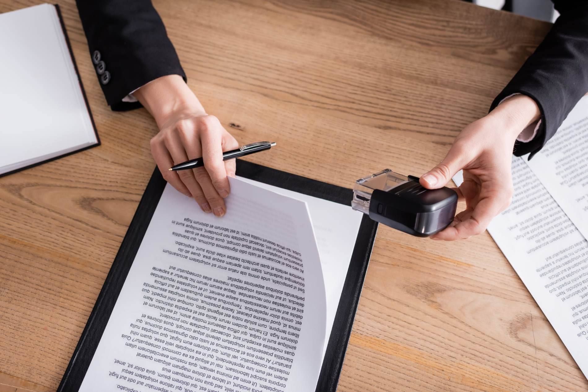 Manos revisando un documento sobre un escritorio con un sello y papeles cerca.