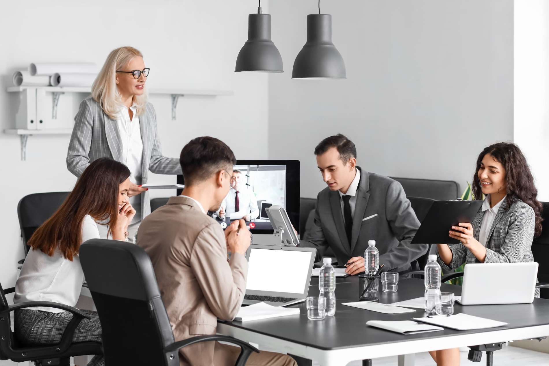 Reunión de equipo en una oficina moderna, con compañeros debatiendo alrededor de una mesa y uno de ellos de pie cerca de un monitor.