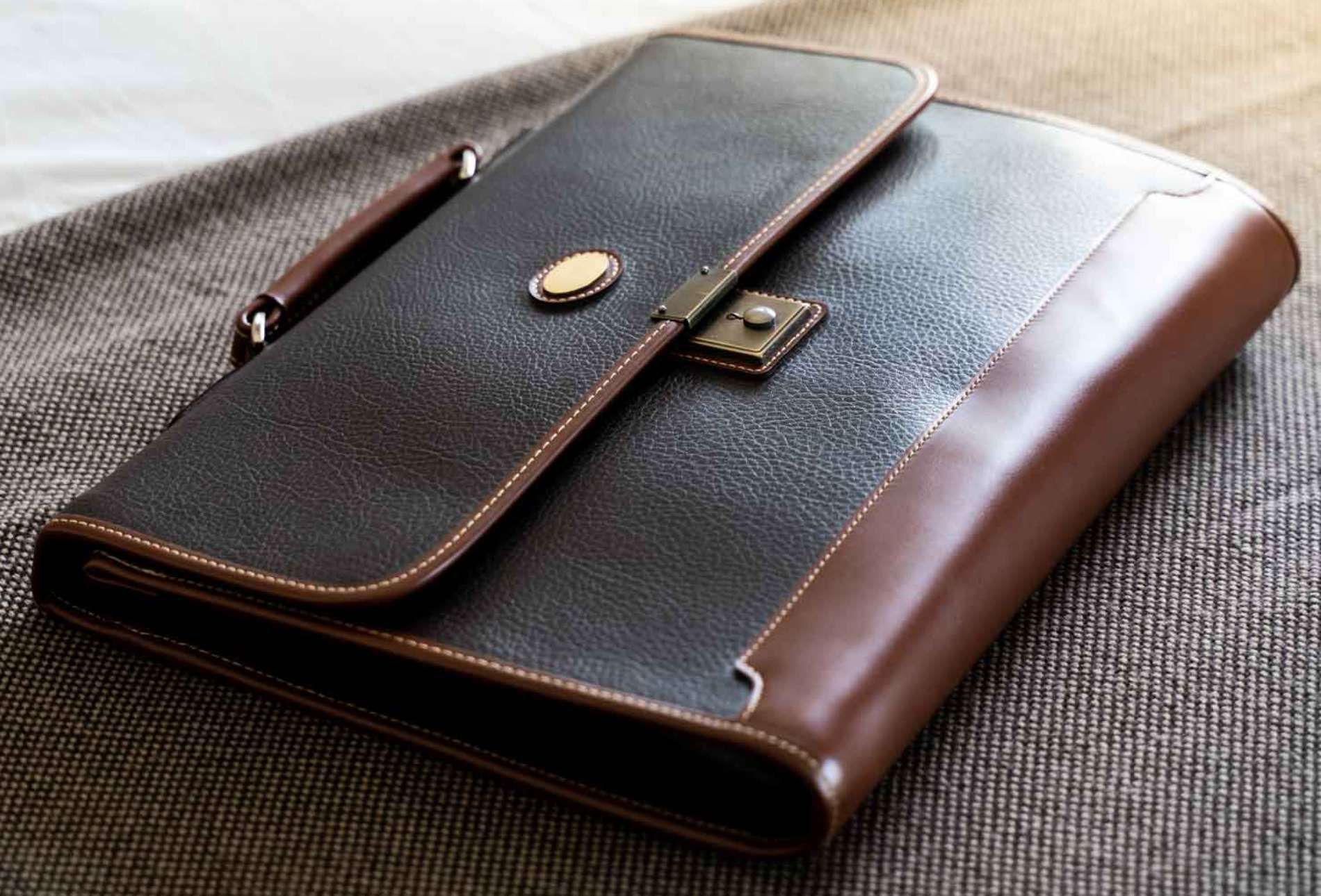 Cartera o estuche de piel negra con ribete marrón y cierre de latón sobre superficie de tela texturizada.
