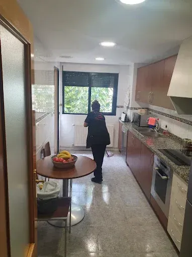 Una persona de pie en una cocina estrecha con armarios marrones, una mesa con fruta y una ventana.