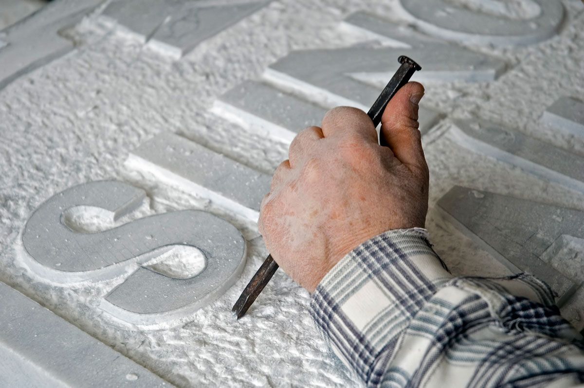 Main ciselant la lettre S dans une surface de pierre blanche, vêtu d'une chemise à carreaux.