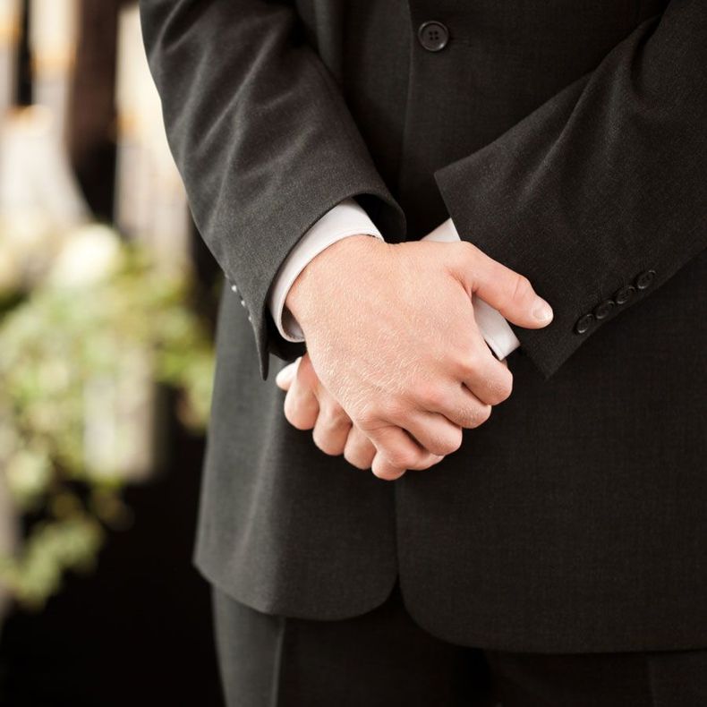 Un homme en costume noir, les mains jointes devant lui, probablement lors d'un enterrement.