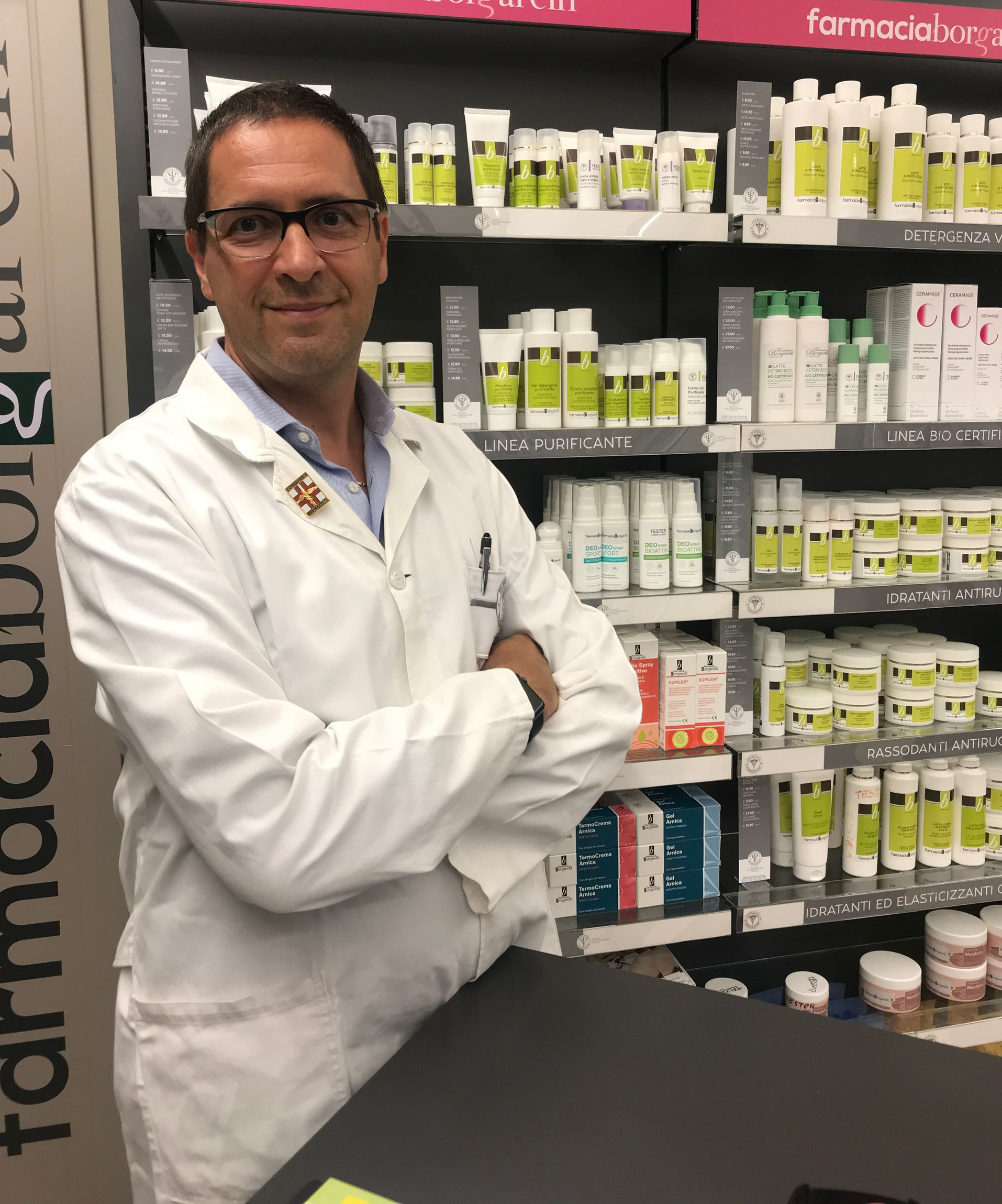 Un farmacista in camice bianco è in piedi in una farmacia, con le braccia incrociate, sorridente. Sullo sfondo, scaffali pieni di prodotti.