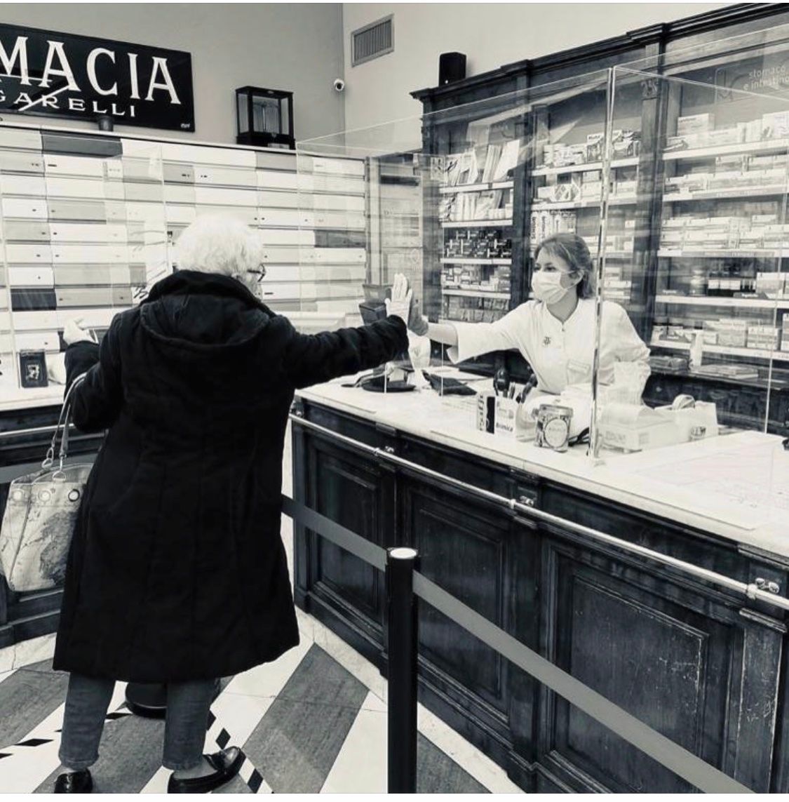 Farmacista con mascherina che consegna i farmaci al cliente al bancone della farmacia, dietro un vetro protettivo.