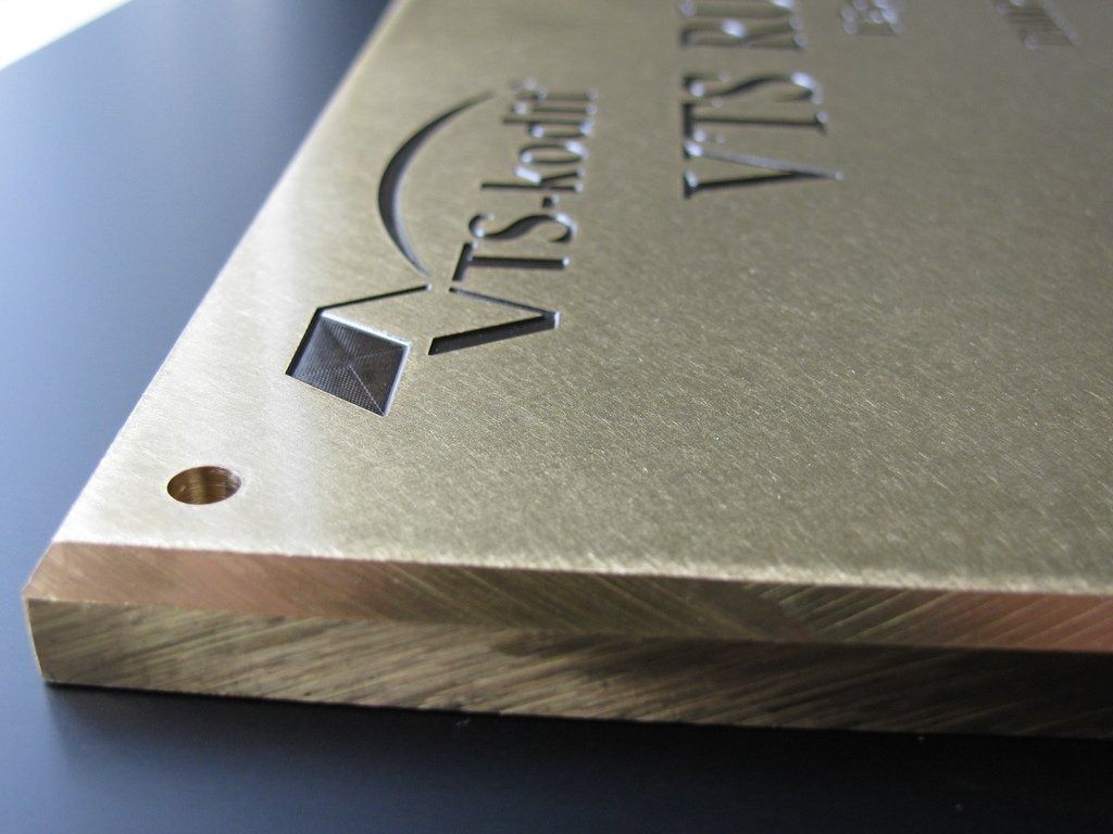 Lähikuva harjatusta metallista laatasta, jossa on kaiverrettu logo ja teksti ja pieni kiinnitysreikä kulmassa