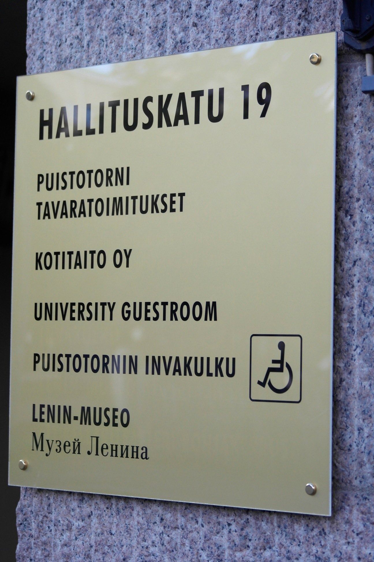 Hallituskatu 19:n kyltti, jossa luetellaan yliopiston vierashuone ja hissi mustalla tekstillä beigenvärisellä laatalla.