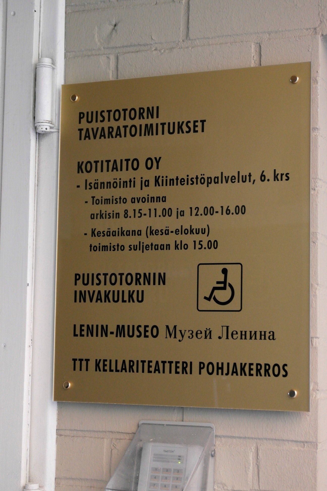 Kultainen seinäkyltti, jossa on suomenkielinen teksti postitoimistolle, hissille, Lenin-museolle ja esteettömälle sisäänkäynnille.