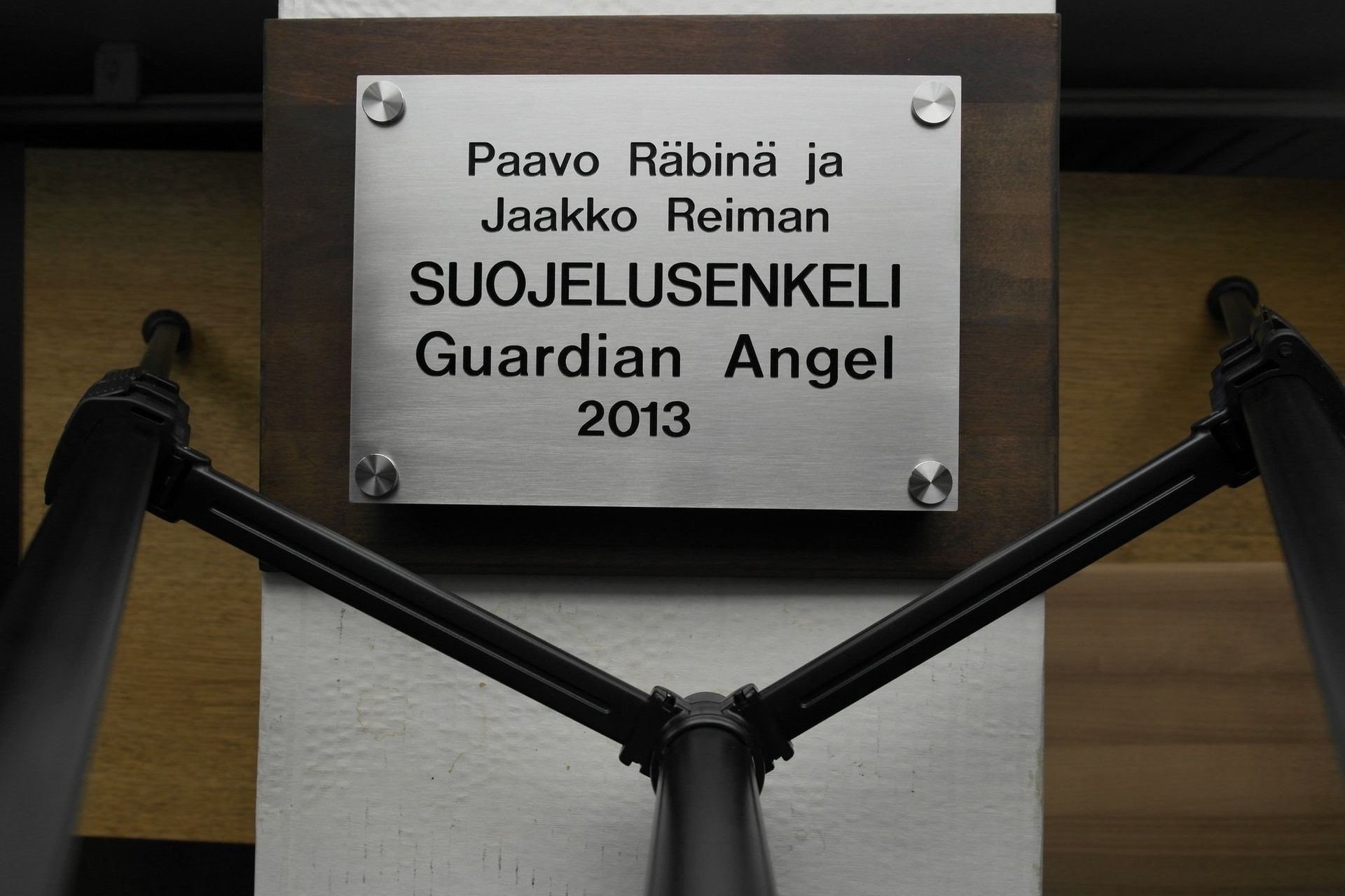 Mustavartiseen puurakenteeseen kiinnitetty laatta, jossa lukee ”Suojelusenkeli / Guardian Angel 2013”.