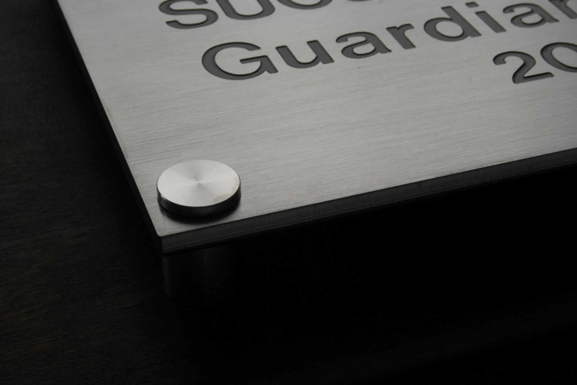 Lähikuva harjatusta metallista laatasta, jossa on kaiverrettu teksti ja hopeinen ruuvi tummalla taustalla