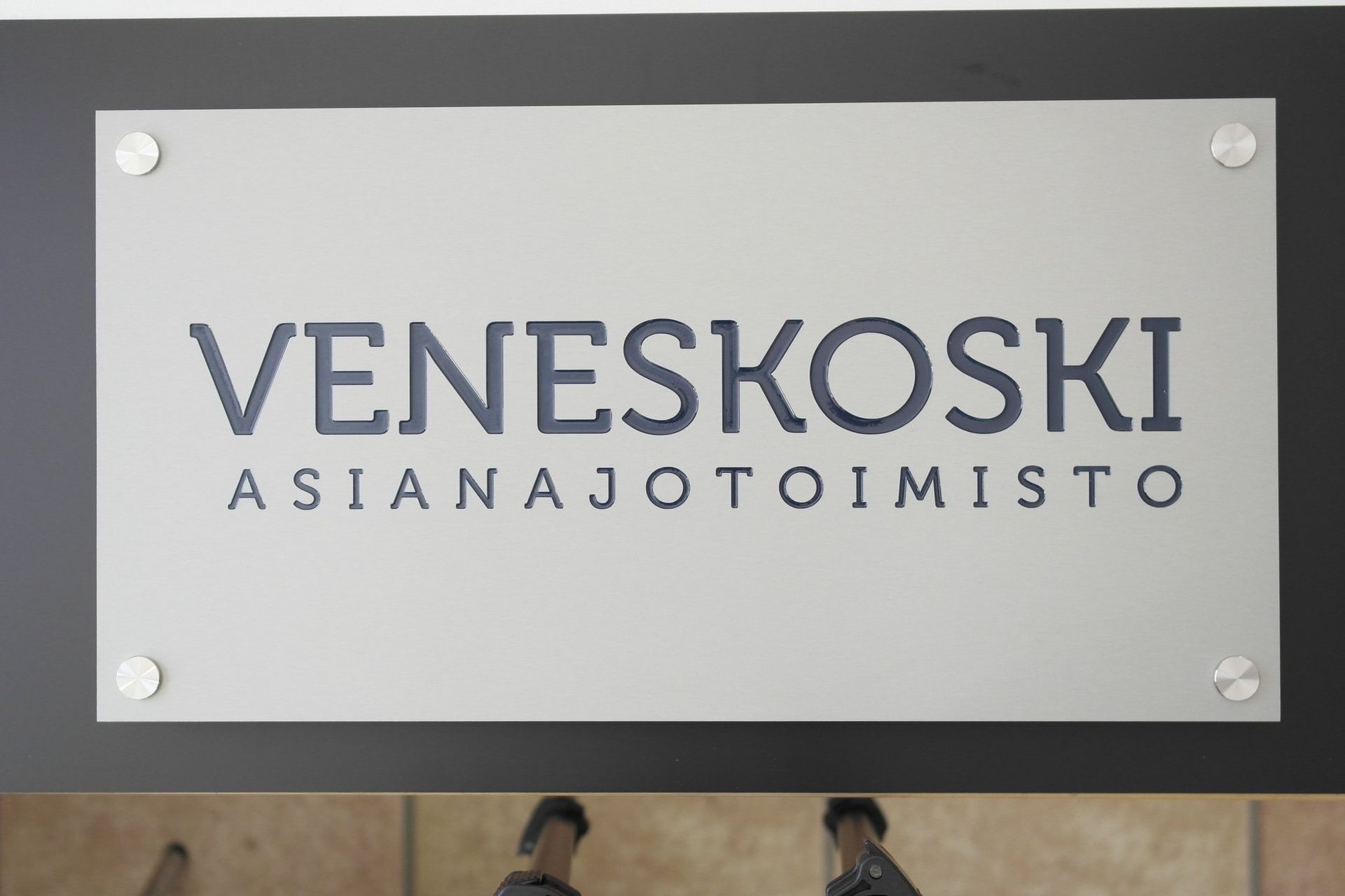 Veneskoski Asianajotoimisto -kyltti hopeisessa seinälaatassa, jossa neljä kulmaruuvia