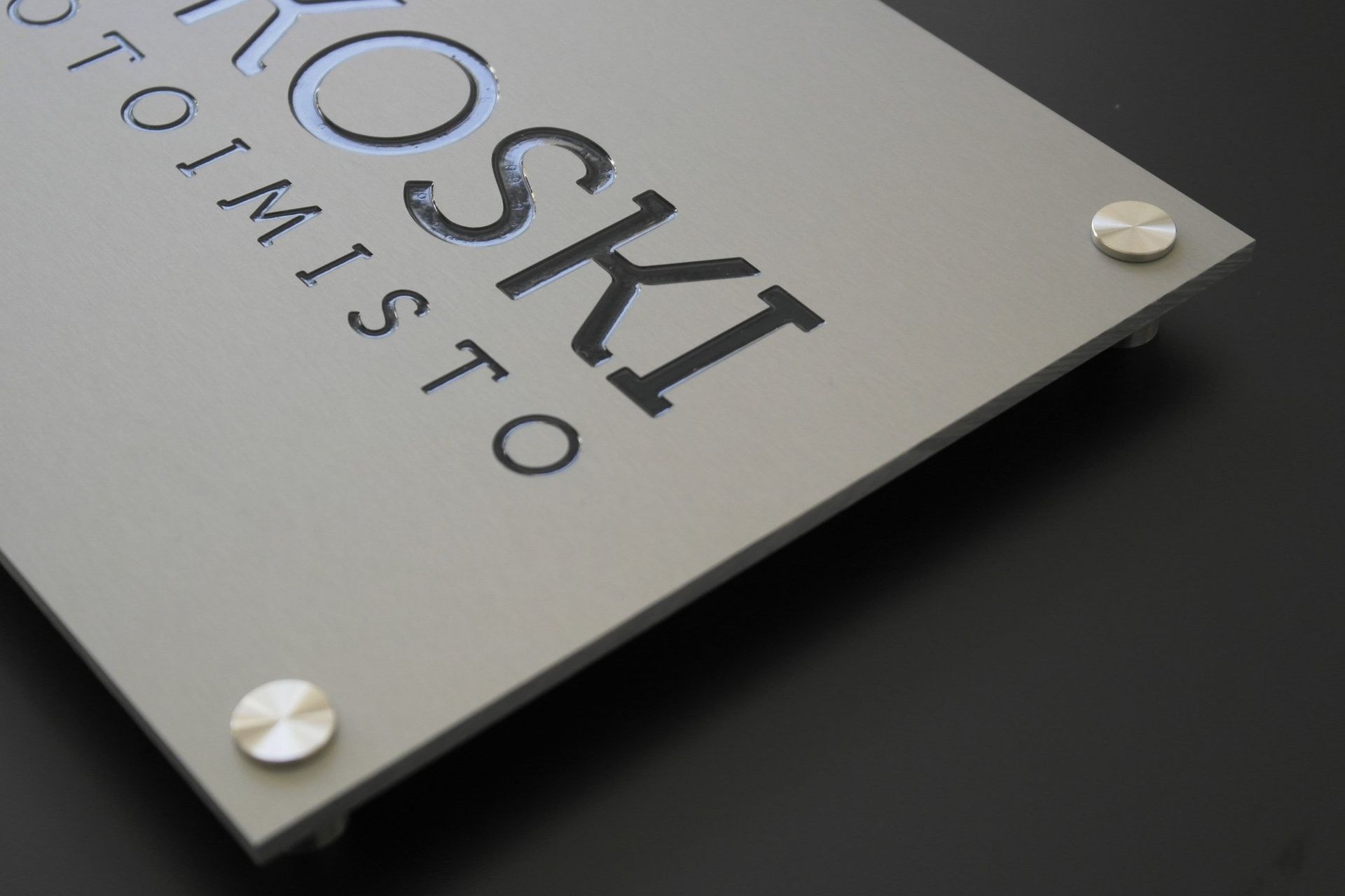 Metallinen nimikyltti, jossa on sana ”Yoshik” ja pienempää tekstiä, kiinnitetty hopearuuveilla tummalle pinnalle