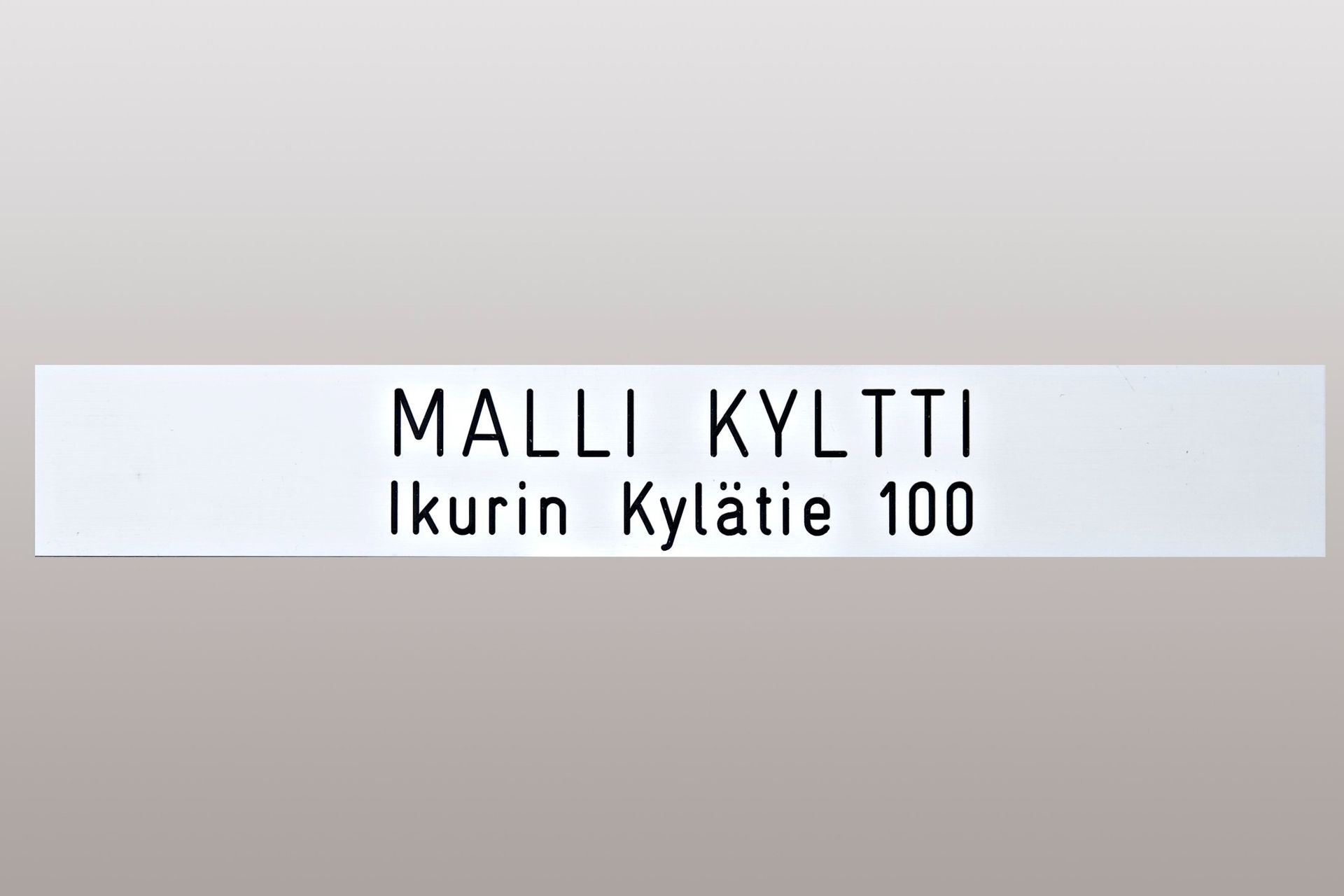 Valkoinen osoitekyltti, jossa lukee ”MALLI KYLTTI” ja ”Ikurin Kylätie 100” harmaalla pohjalla