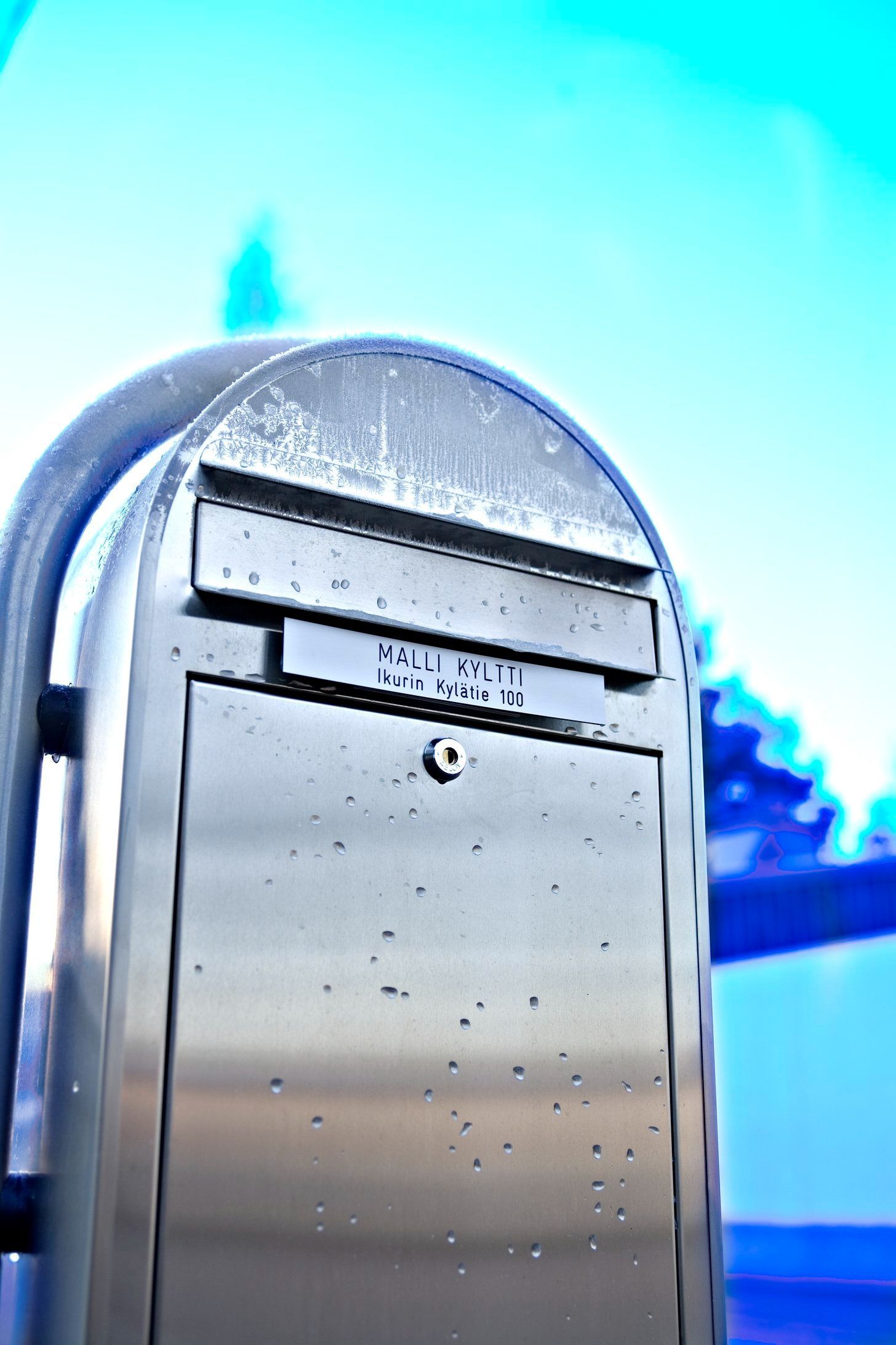 Ruostumattomasta teräksestä valmistettu postilaatikko kirkkaan sinistä taivasta vasten, taustalla talon katto.