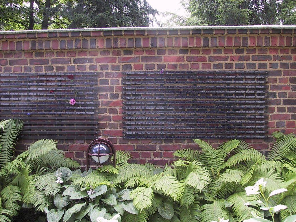 Tiilinen muistomuuri, jossa kaksi tummaa laattaa saniaisten ja vihreyden yläpuolella ulkona