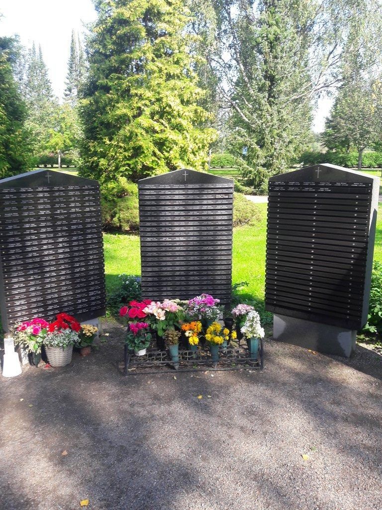 Kolme mustaa muistolaattaa värikkäillä kukkakimpuilla aurinkoisella puutarhamuistoalueella