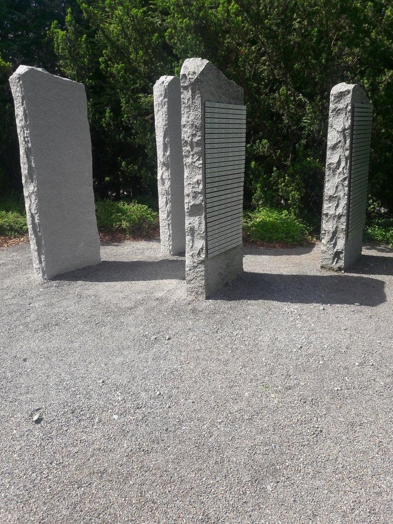 Kolme harmaata kiviveistosta seisoo sora-alueella ulkona puiden taustalla