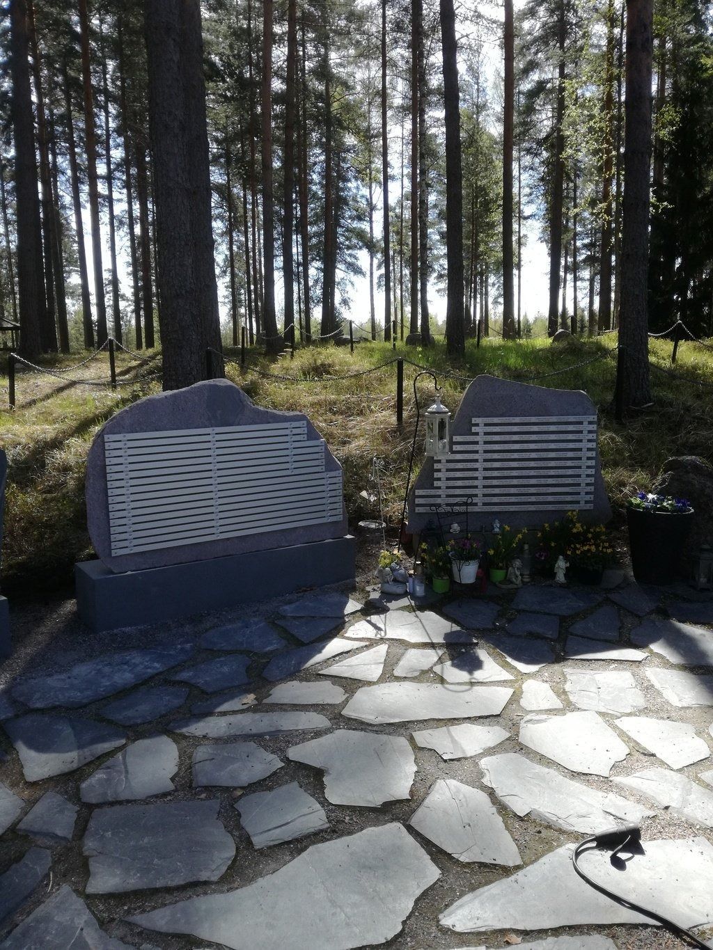 Kaksi valkoista muistolaattaa kivipenkillä mäntymetsässä, ja niiden juurella on kukkia.