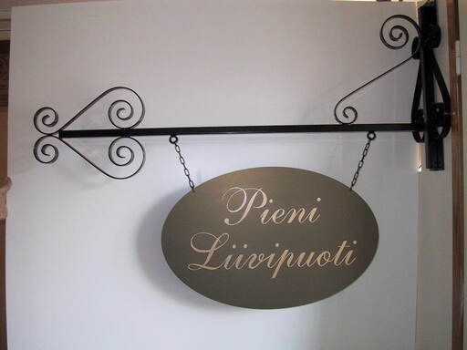 Soikea riippuva kyltti, jossa lukee ”Pieni Liivipuoti” mustassa koristeellisessa seinäkiinnikkeessä