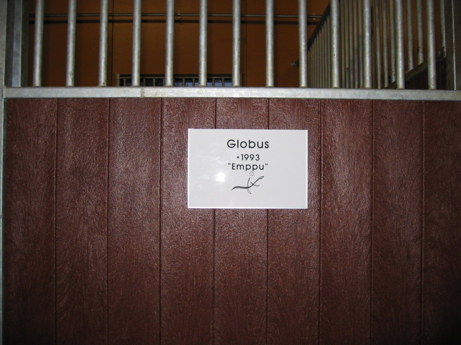 Valkoinen kyltti ruskealla tallin ovella, jossa teksti ja hevosen luonnos metallitankojen alla.