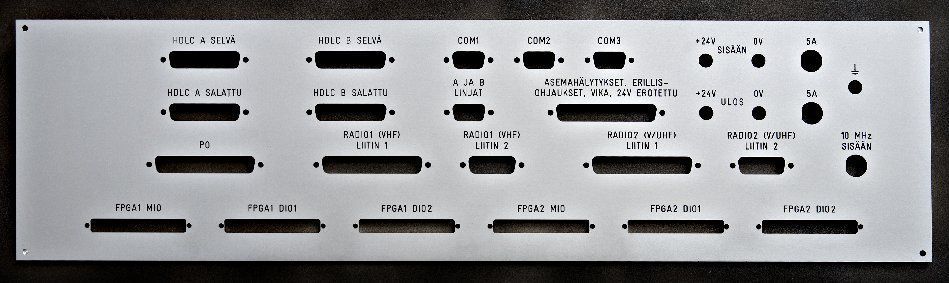 Valkoinen elektroninen ohjauspaneeli, jossa on useita mustia painikkeita, merkkivaloja ja merkittyjä kytkimiä metallipinnalla