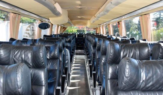 Interior de un autobús vacío con filas de asientos de cuero negro, compartimentos superiores para equipaje y ventanas laterales.