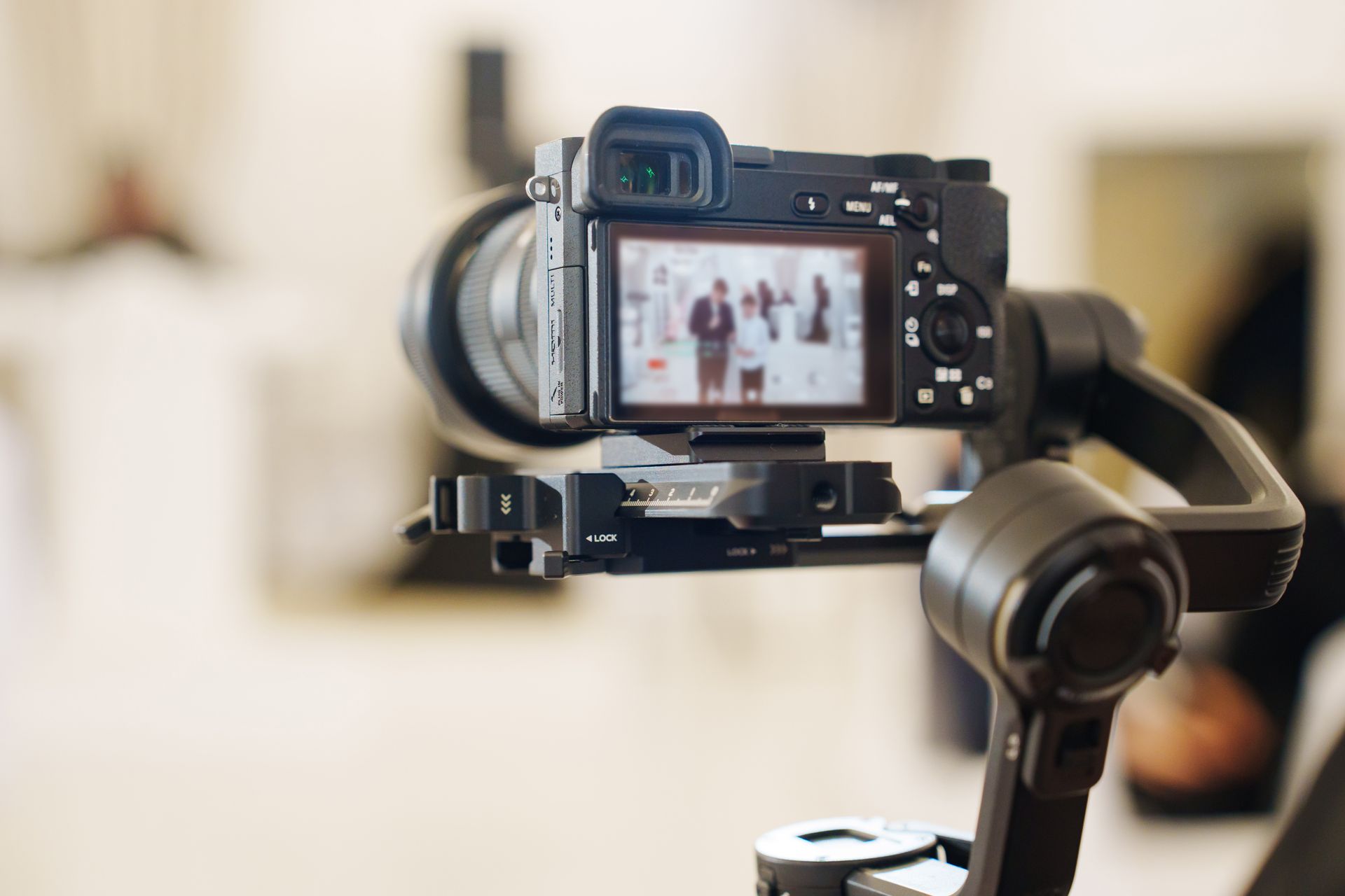 Schwarze Kamera auf einem Gimbal, Bildschirm zeigt Personen. Innenaufnahme mit unscharfem Hintergrund.