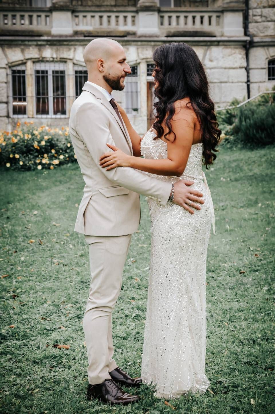 Ein Paar in festlicher Kleidung umarmt sich lächelnd in einem Garten. Beigefarbener Anzug, weisses Paillettenkleid, im Hintergrund ein Steingebäude.