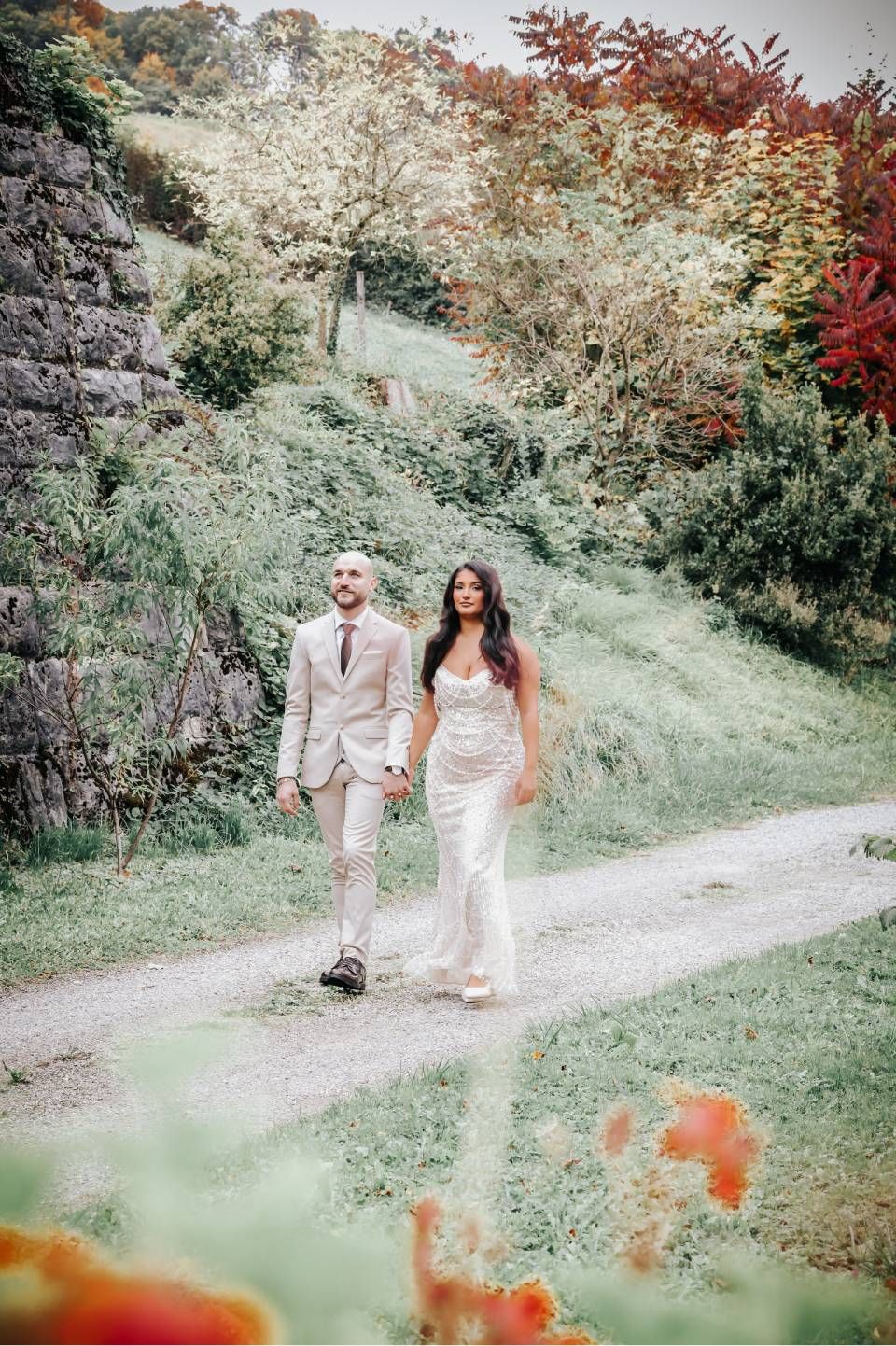 Ein Paar in Hochzeitskleidung spaziert Hand in Hand auf einem Pfad durch eine üppig grüne Landschaft.