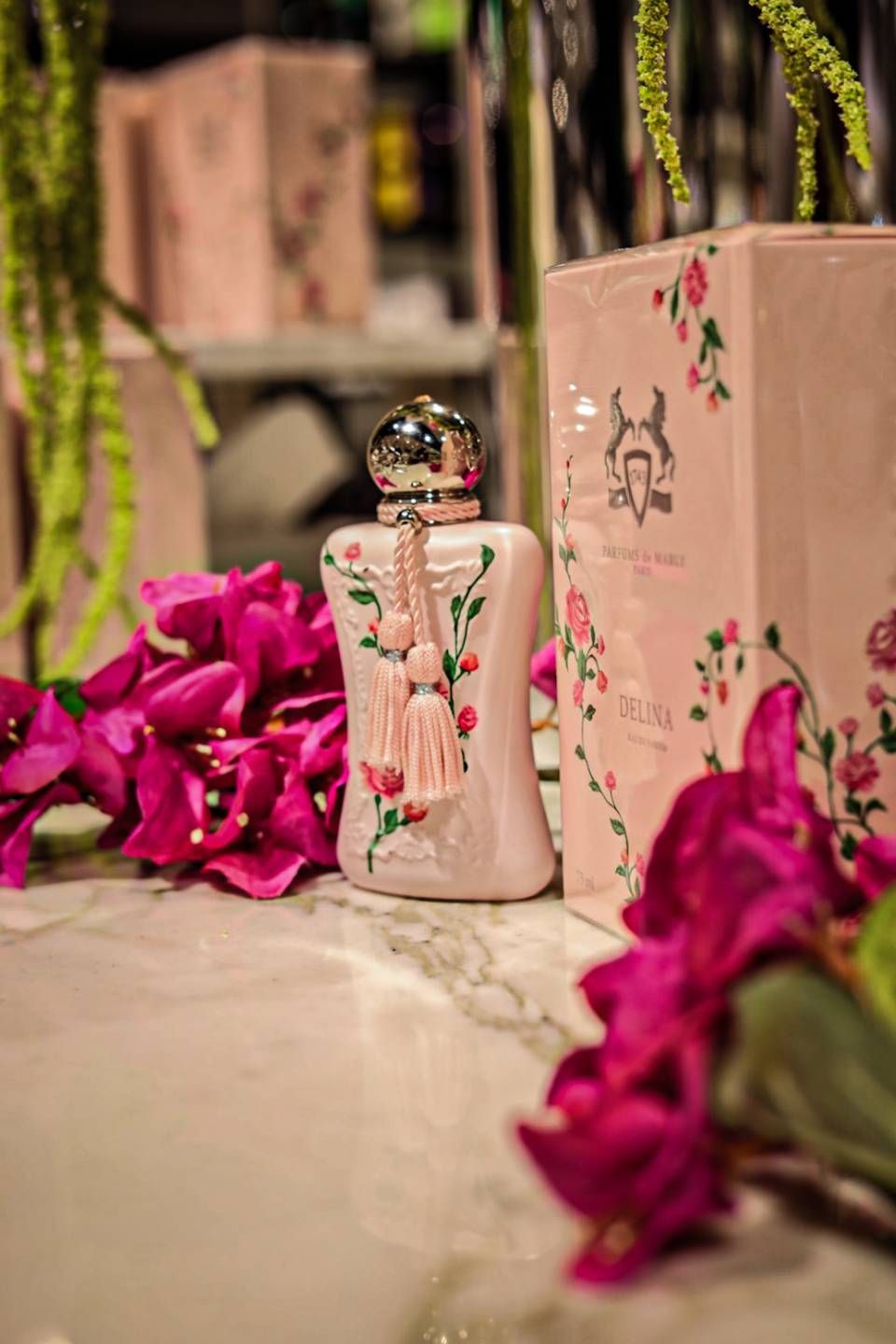 Rosa Parfümflakon mit floralem Muster, umgeben von rosa Blüten und Verpackung auf einer Marmoroberfläche.