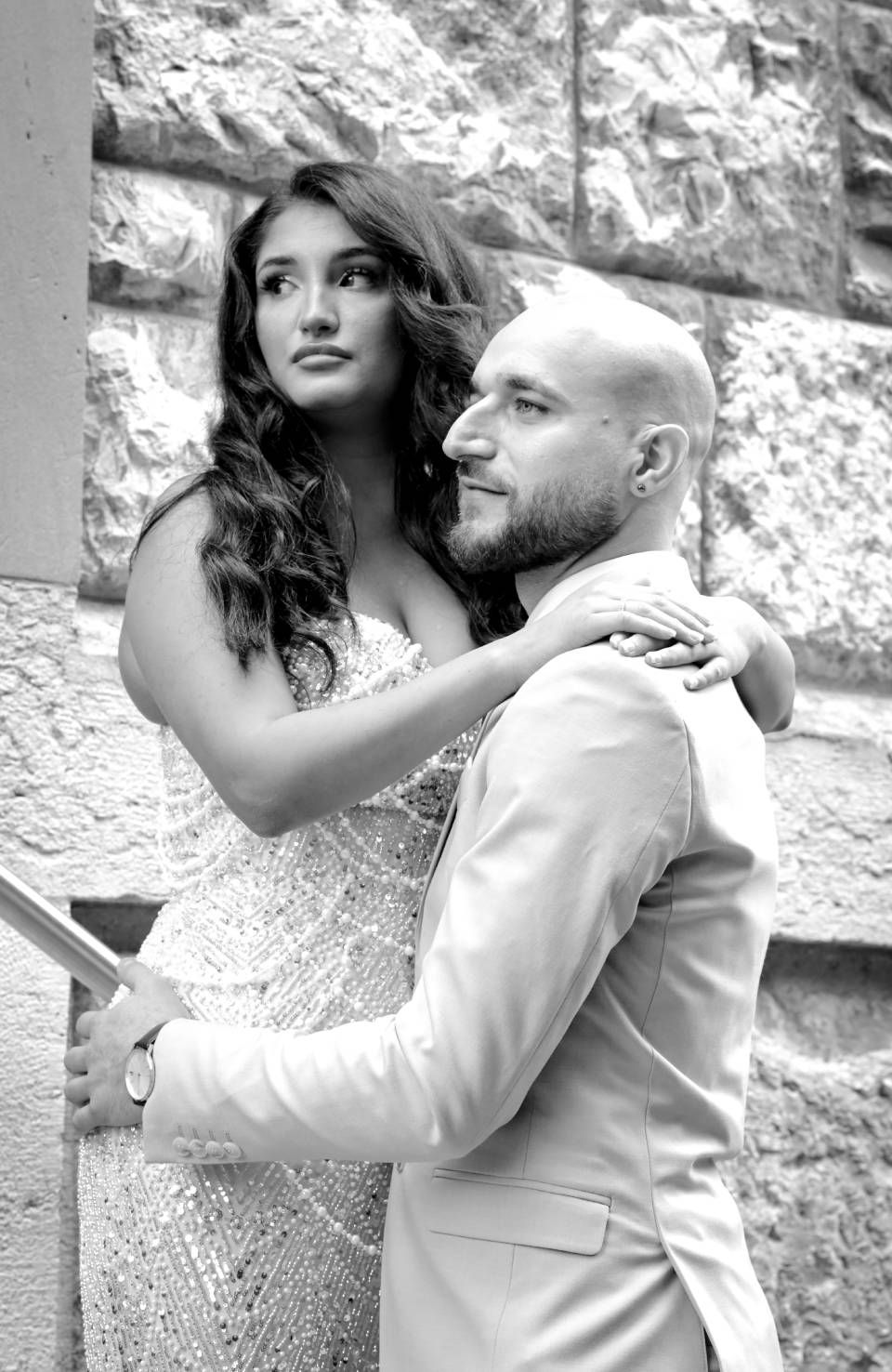 Frau in funkelndem Kleid umarmt Mann im Anzug im Freien, in der Nähe einer Steinmauer.