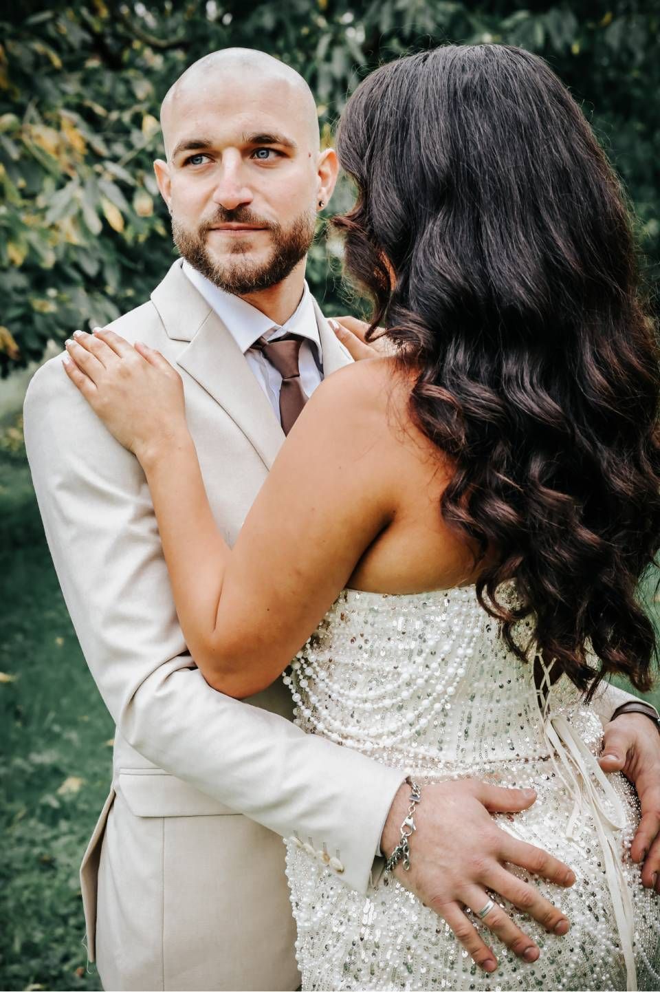 Ein Mann im beigen Anzug umarmt im Freien eine Frau in einem glitzernden weissen Kleid.