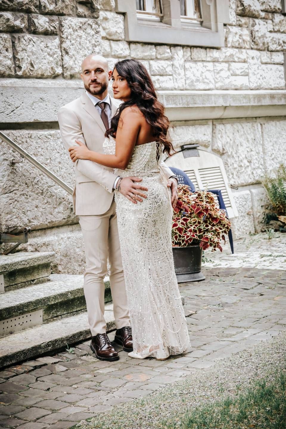 Ein Paar in festlicher Kleidung umarmt sich im Freien vor einem Steingebäude. Die Frau trägt ein besticktes Kleid, der Mann einen beigen Anzug.