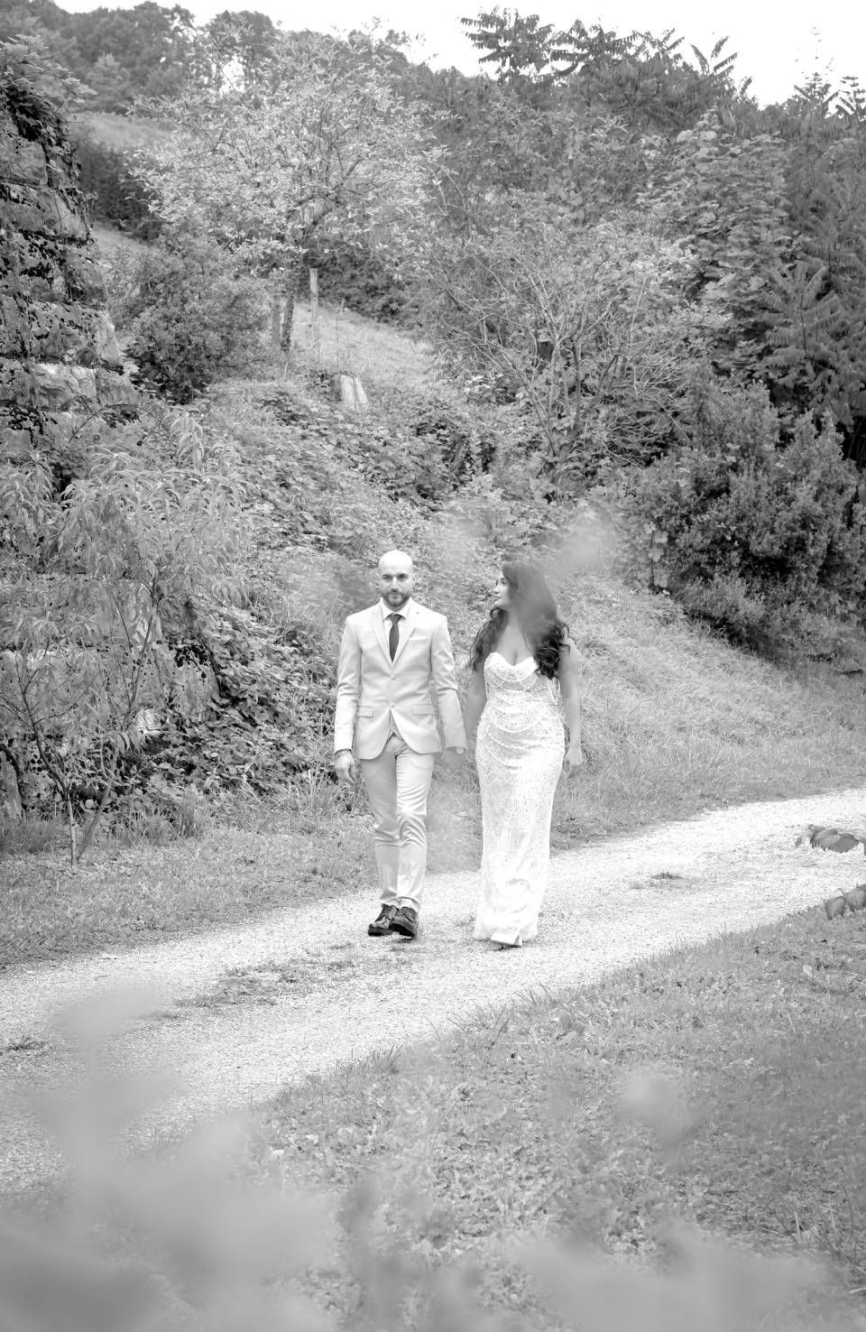 Ein Paar spaziert Hand in Hand auf einem Pfad, umgeben von Bäumen und Grün.