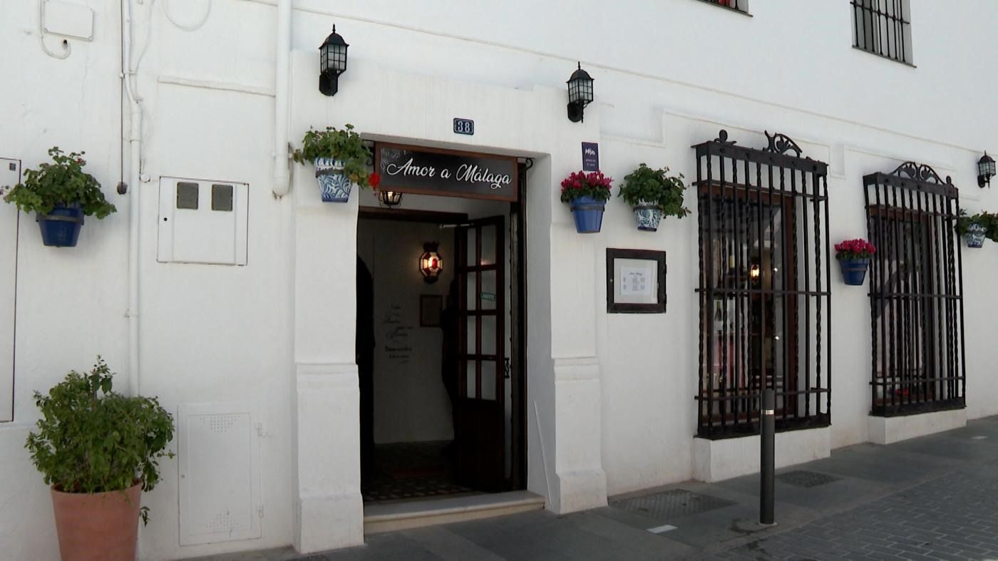 Fachada blanca con puertas y ventanas de hierro negro, plantas en macetas y faroles colgantes en la acera.