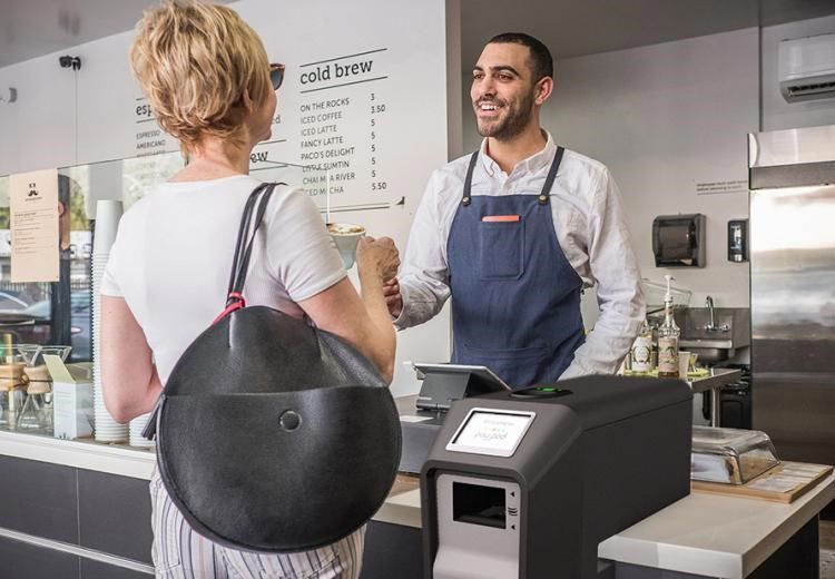 Un cliente y un barista charlan en la barra de una cafetería, con la caja registradora y la carta detrás de ellos.