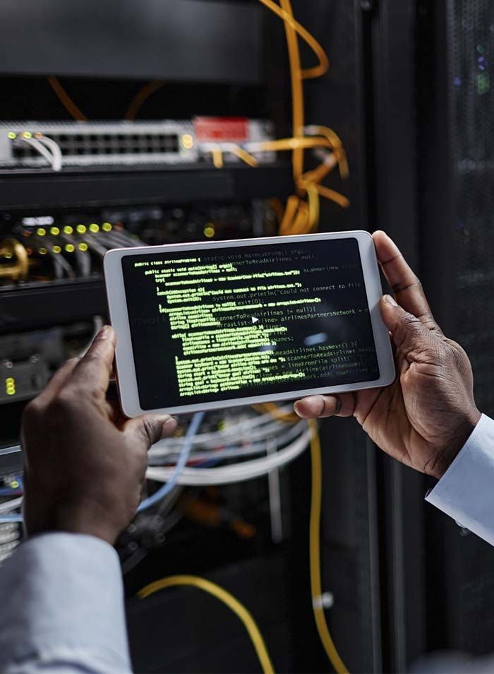 Manos sosteniendo una tableta con un código verde en una sala de servidores, con cables de red y equipos detrás.