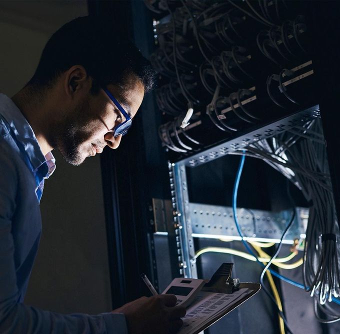 Técnico inspeccionando cables de red en un rack de servidores mientras usa una tableta, en un centro de datos con poca luz.