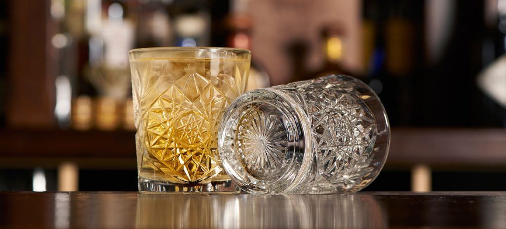 Un verre plein de whisky ambré repose à côté d'un verre en cristal décoratif vide sur un comptoir de bar en bois poli.