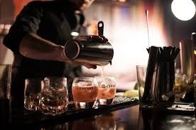Un barman verse un cocktail d'un shaker en métal dans des verres posés sur un comptoir faiblement éclairé.