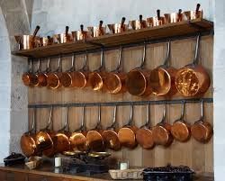 Deux rangées horizontales de casseroles et de poêles en cuivre poli sont suspendues à des supports fixés à un mur en bois.