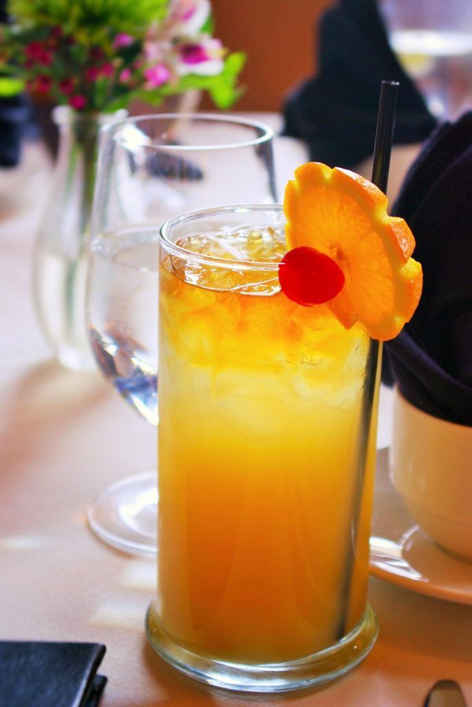 Un grand cocktail jaune, garni d'une tranche d'orange et d'une cerise, posé sur une table au fond flou.