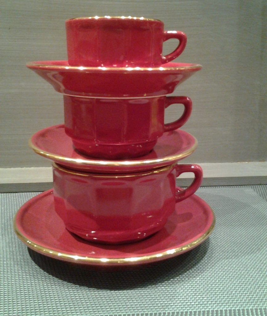 Une pile de trois tasses et soucoupes en céramique rouge à bord doré, disposées verticalement sur une surface texturée.